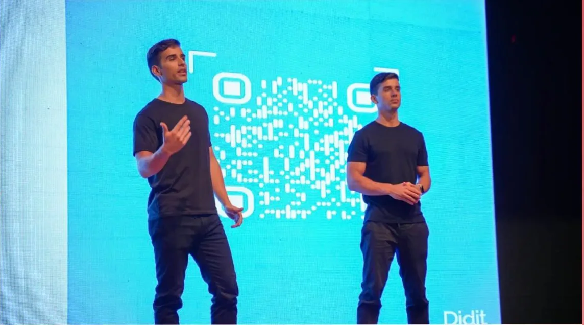 Alejandro and Alberto on stage in black t-shirts in front of a large blue screen with a QR code
