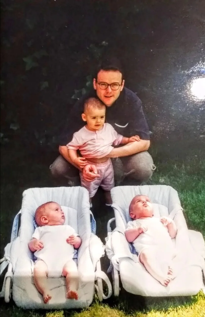 A man in glasses crouching with a toddler, in front of two newborn twins in matching baby bouncers on the grass
