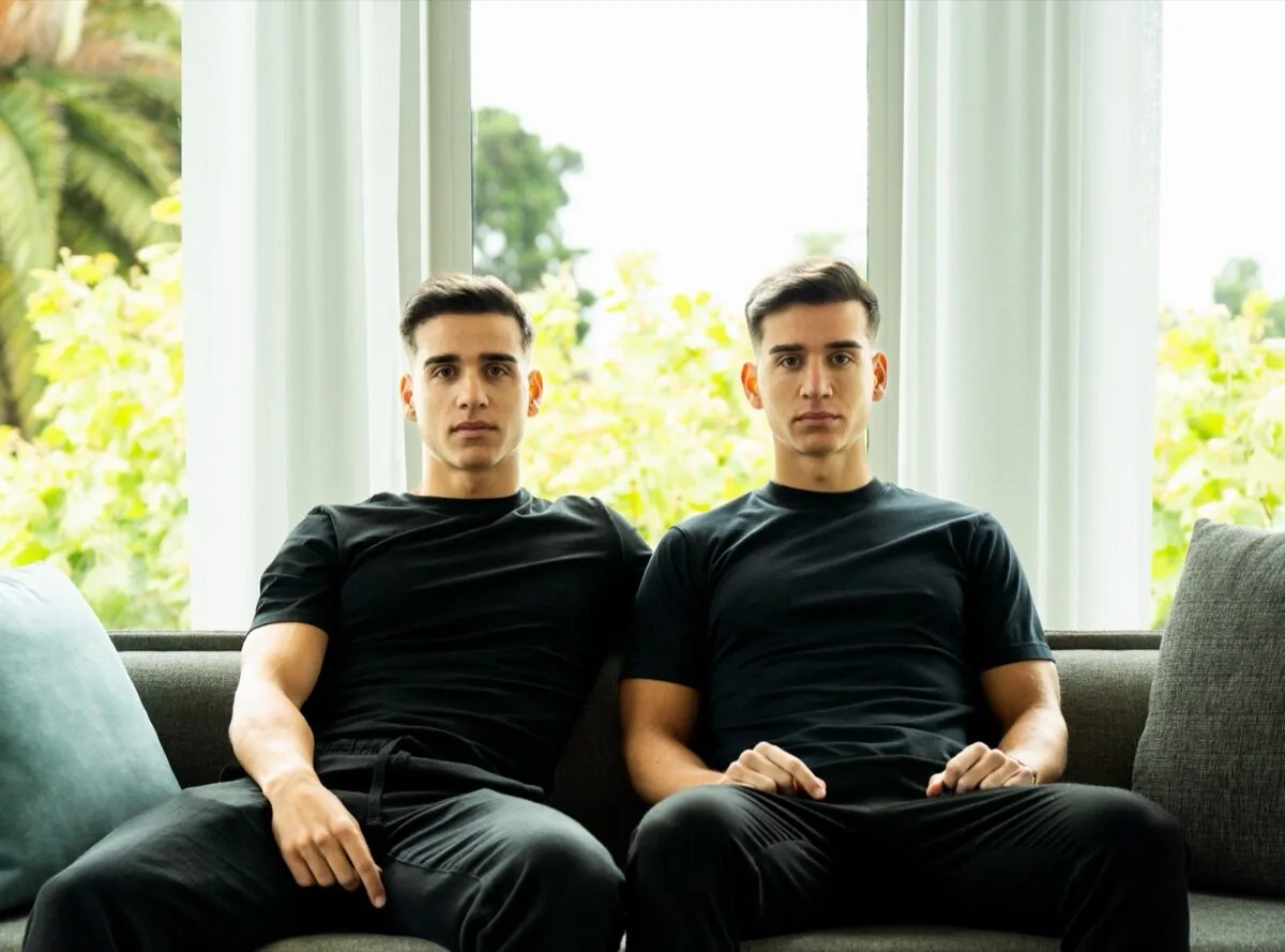 The twins sitting side by side on a sofa, in black t-shirts, same pose